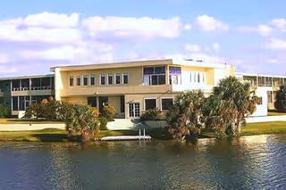 Exterior view of a two-story assisted living residence building named Oasis at Margate, situated beside a body of water with palm trees and a clear sky with some clouds.