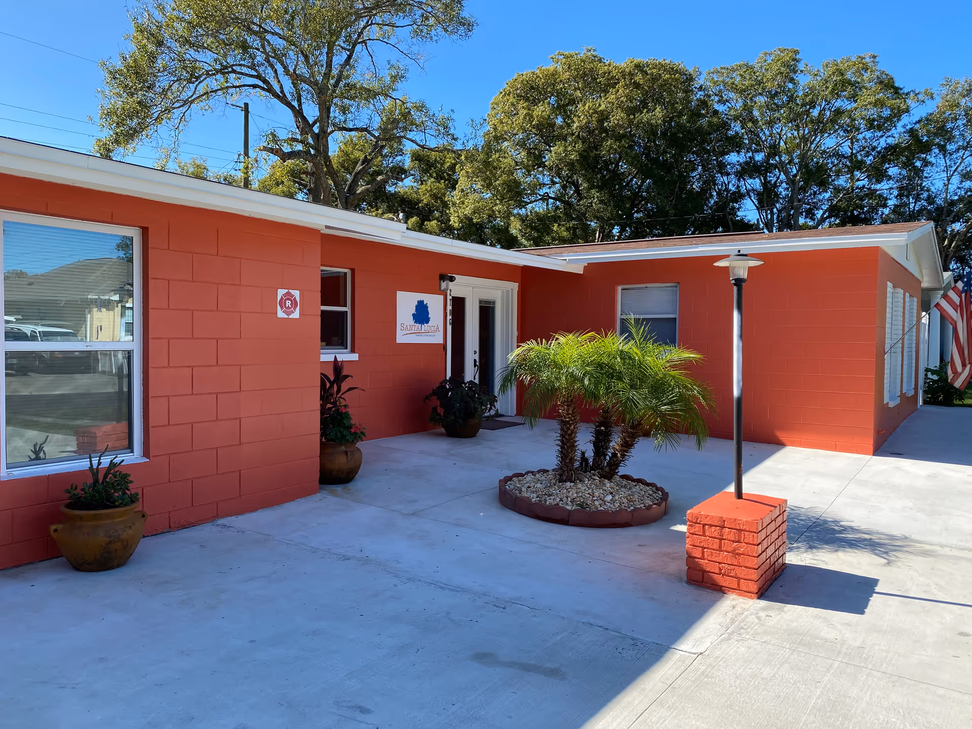 Exterior view of a single-story red building with white trim, featuring a small landscaped area with palm plants in the center of a concrete courtyard. There are potted plants near the building walls, a lamp post on a red brick base, and an American flag visible on the right side. The building has windows and a door with a sign that reads 'Santa Lucia ALF Inc'. Trees and a clear blue sky are in the background.