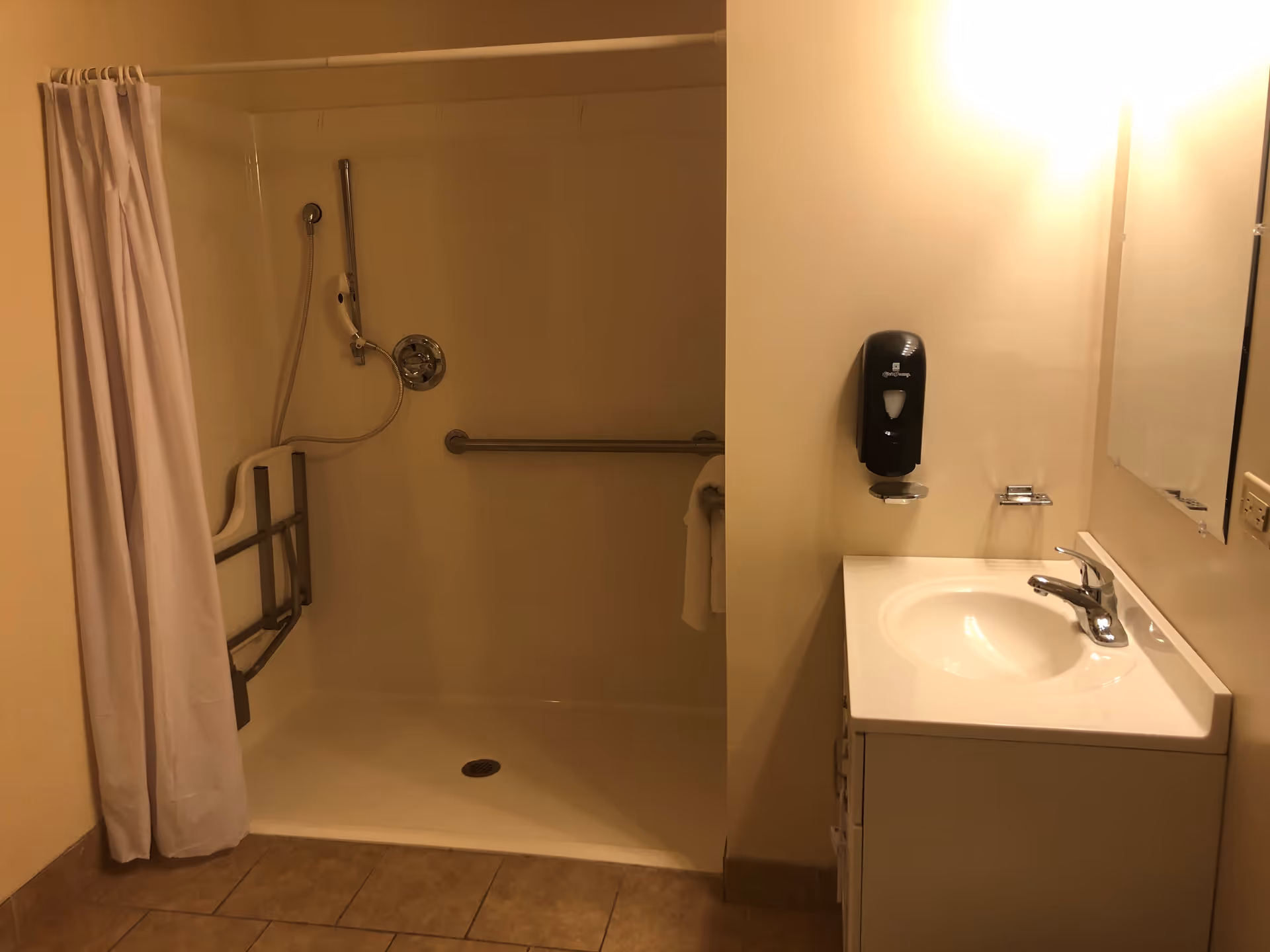 A bathroom with a walk-in shower featuring a white curtain, a handheld showerhead, and grab bars for accessibility. To the right is a white sink with a faucet, a mounted soap dispenser, and a mirror above the sink. The floor is tiled in a light brown color.