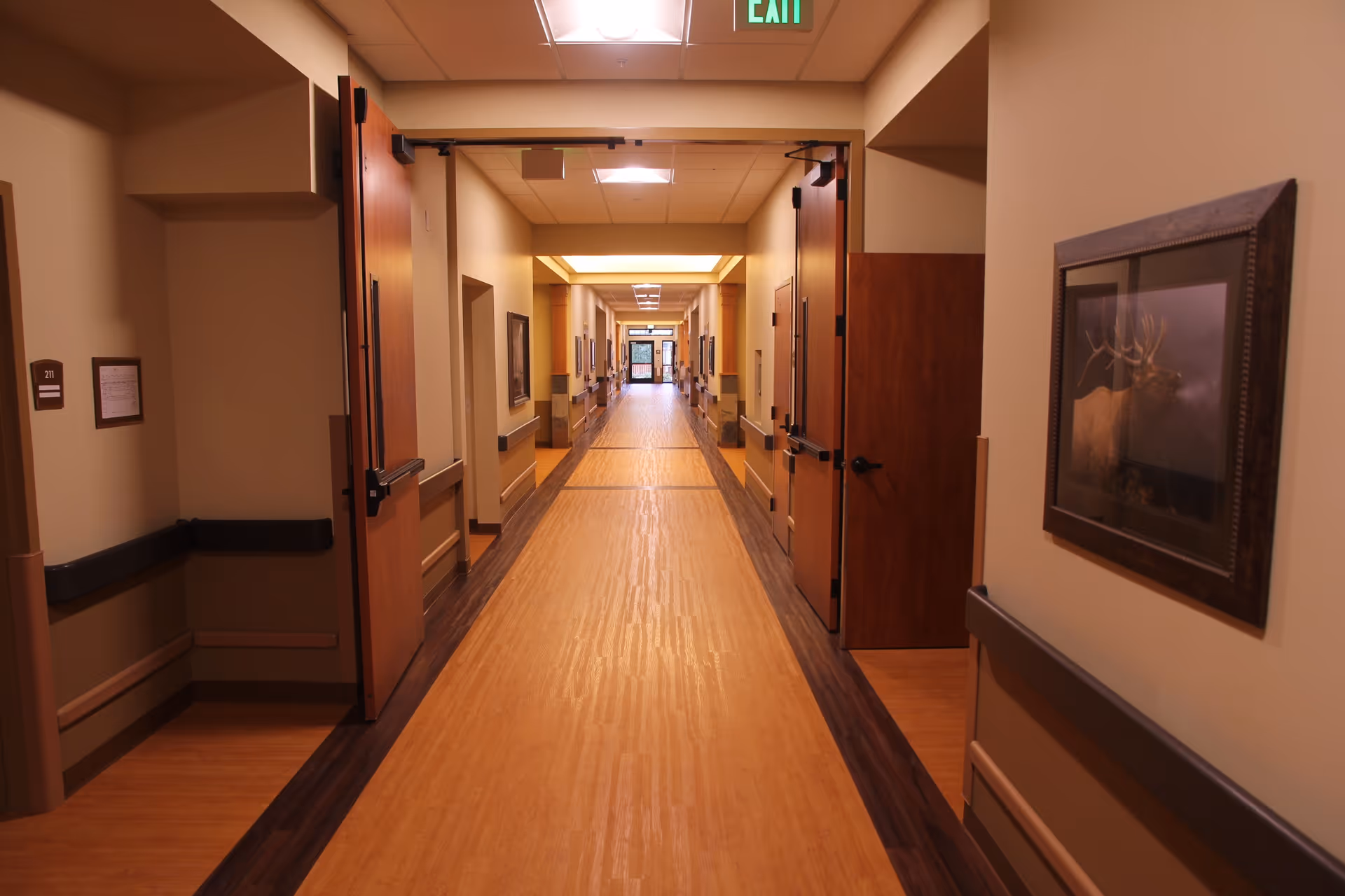 A long, well-lit hallway in a senior living facility with wooden floors and beige walls. Several open wooden doors line the corridor, and there is a framed picture of a deer on the right wall. An exit sign is visible on the ceiling near the foreground.