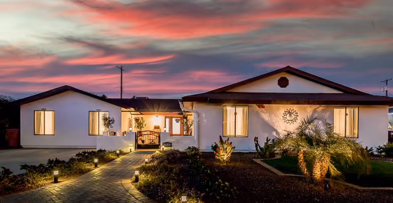 Exterior view of a single-story assisted living facility building at dusk with a paved walkway lined with small lights leading to the entrance. The sky is colorful with pink and purple hues, and the building is white with a brown roof and several windows illuminated from inside. There are plants and landscaping around the walkway and building.