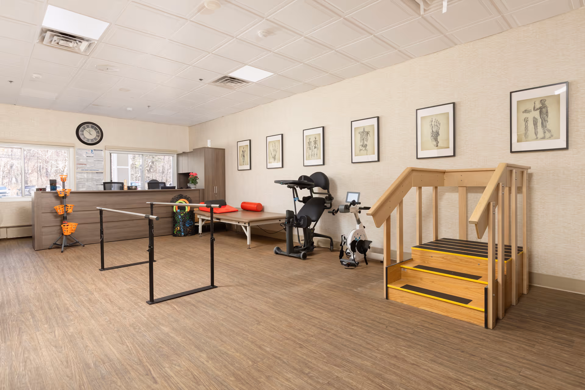 A physical therapy or rehabilitation room with wooden flooring and beige walls. The room contains parallel bars for walking exercises, a stationary exercise bike, a padded therapy table with red bolster cushions, and a small wooden staircase with handrails for step training. There is a reception desk with office chairs and a clock on the wall above two windows. Several framed anatomical drawings are hung on the wall.