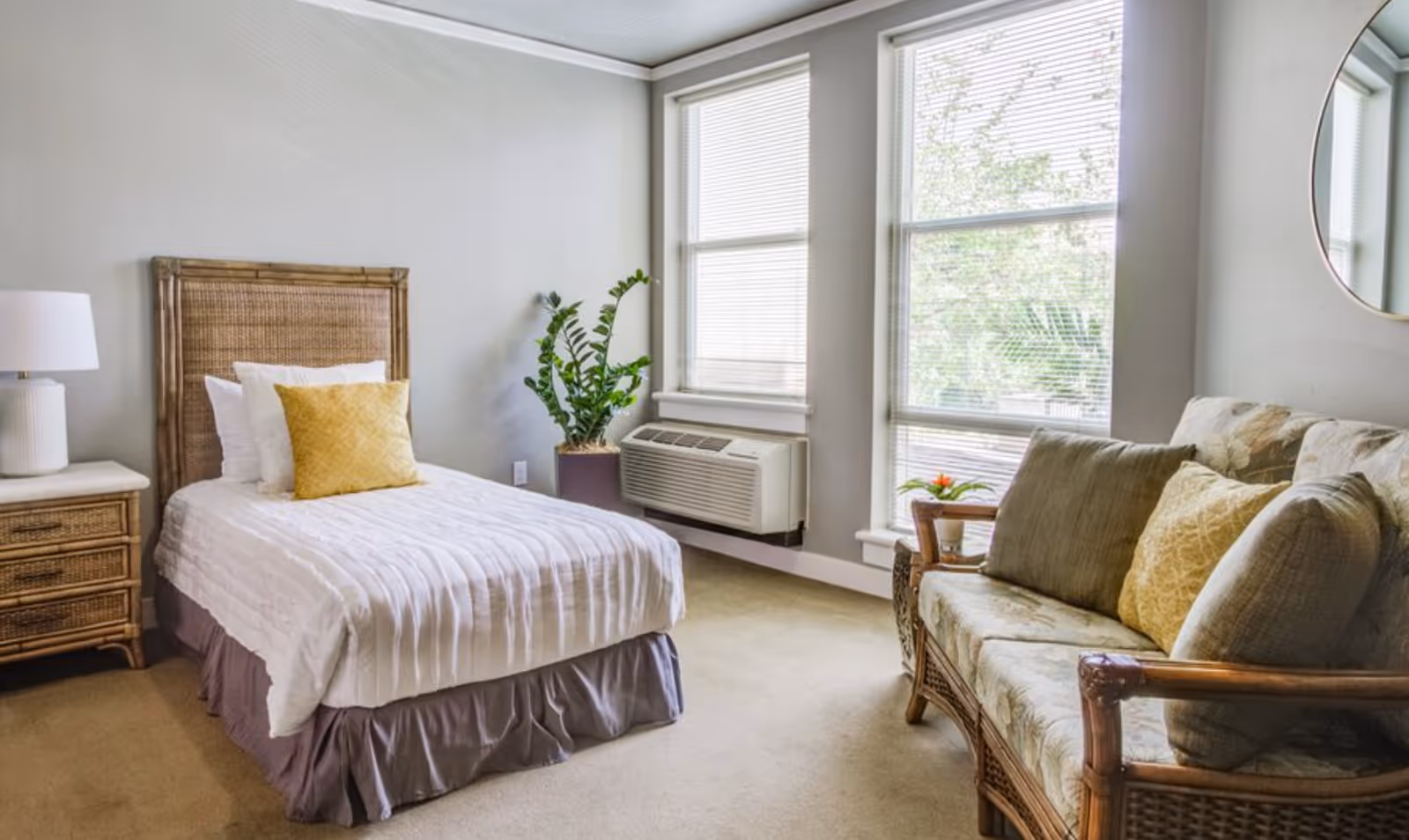 Bright furnished studio-style bedroom with a single bed, wicker nightstand, potted plant, window air unit, and a sofa.