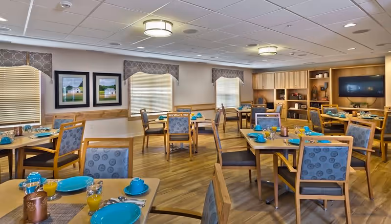 A bright dining room with multiple wooden tables and chairs arranged neatly. Each table is set with turquoise plates, cups, and glasses filled with orange juice. The room has wooden flooring, three windows with blinds and patterned valances, framed artwork on the walls, and built-in wooden cabinetry with a mounted flat-screen TV.