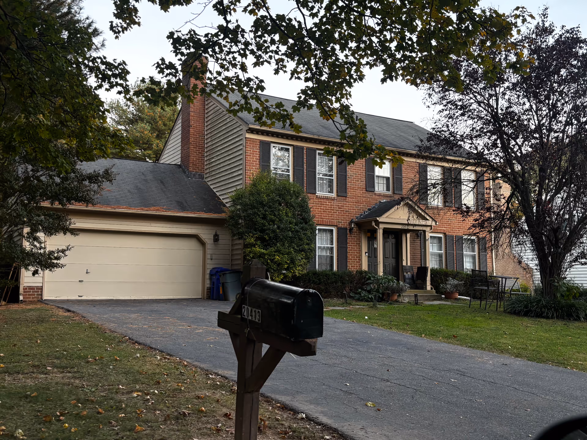 A two-story brick house with black shutters and a black front door under a small porch. There is a driveway leading to a beige garage door on the left side. A black mailbox with the number 20413 is in the foreground. The yard has green grass, some bushes, and trees with leaves partially framing the image.