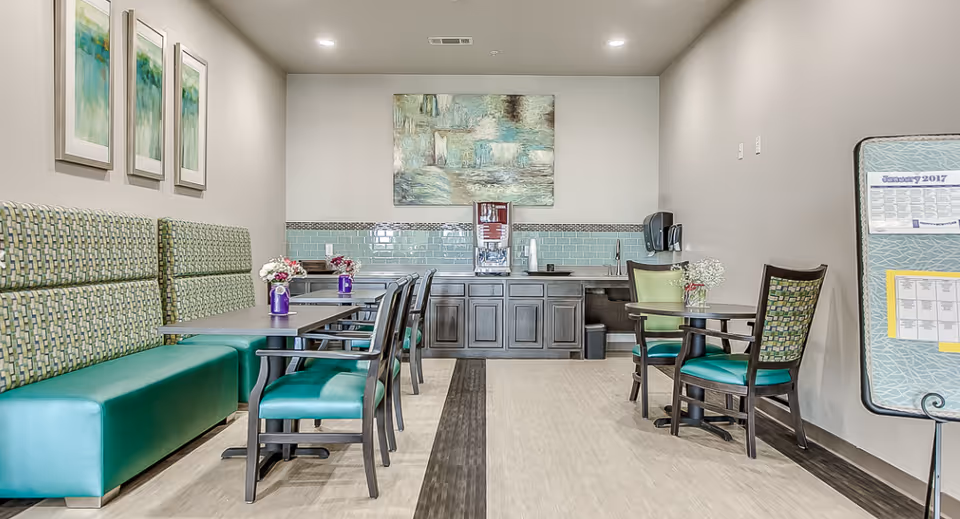 Bright communal dining area with green booth seating, tables and chairs and a counter with a beverage dispenser.