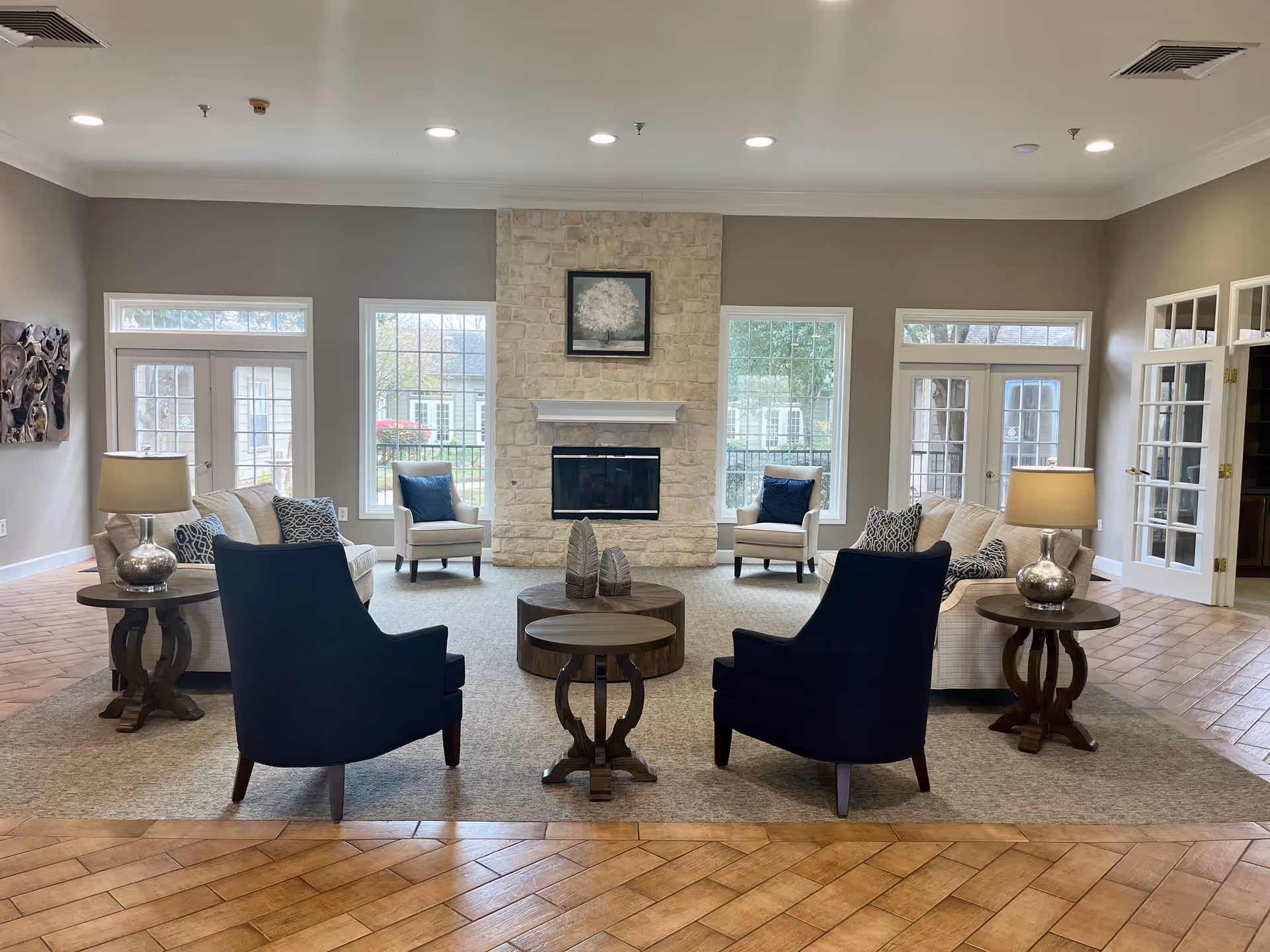 A spacious and well-lit senior living facility common area with a stone fireplace centered on the far wall. The room features two beige sofas with patterned pillows, two beige armchairs with dark blue pillows, and two dark blue armchairs arranged around a round wooden coffee table and two matching side tables with lamps. Large windows and glass doors let in natural light, and the floor is a combination of wood and carpet.