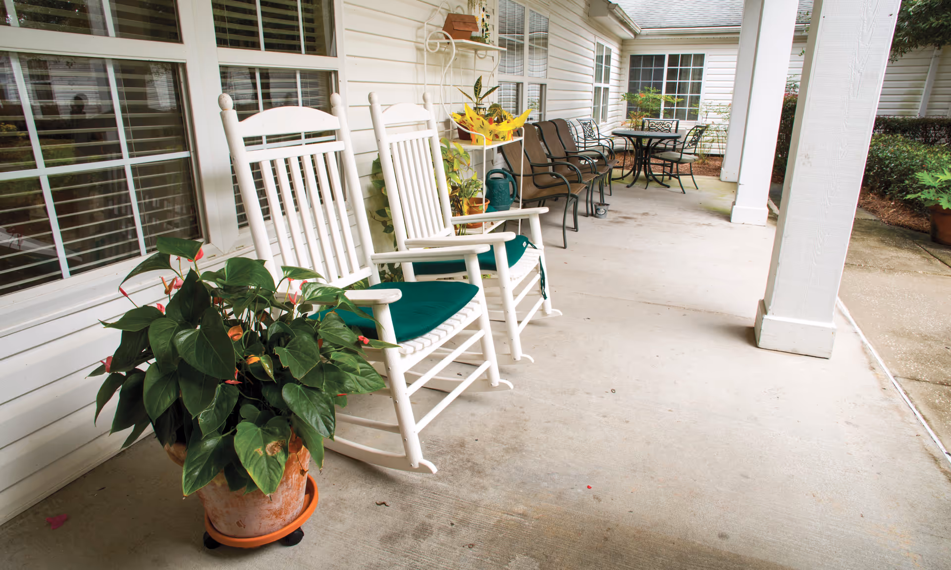 Covered outdoor patio area with white rocking chairs with green cushions, potted plants, metal chairs, and a round metal table. The patio is adjacent to a white building with large windows.