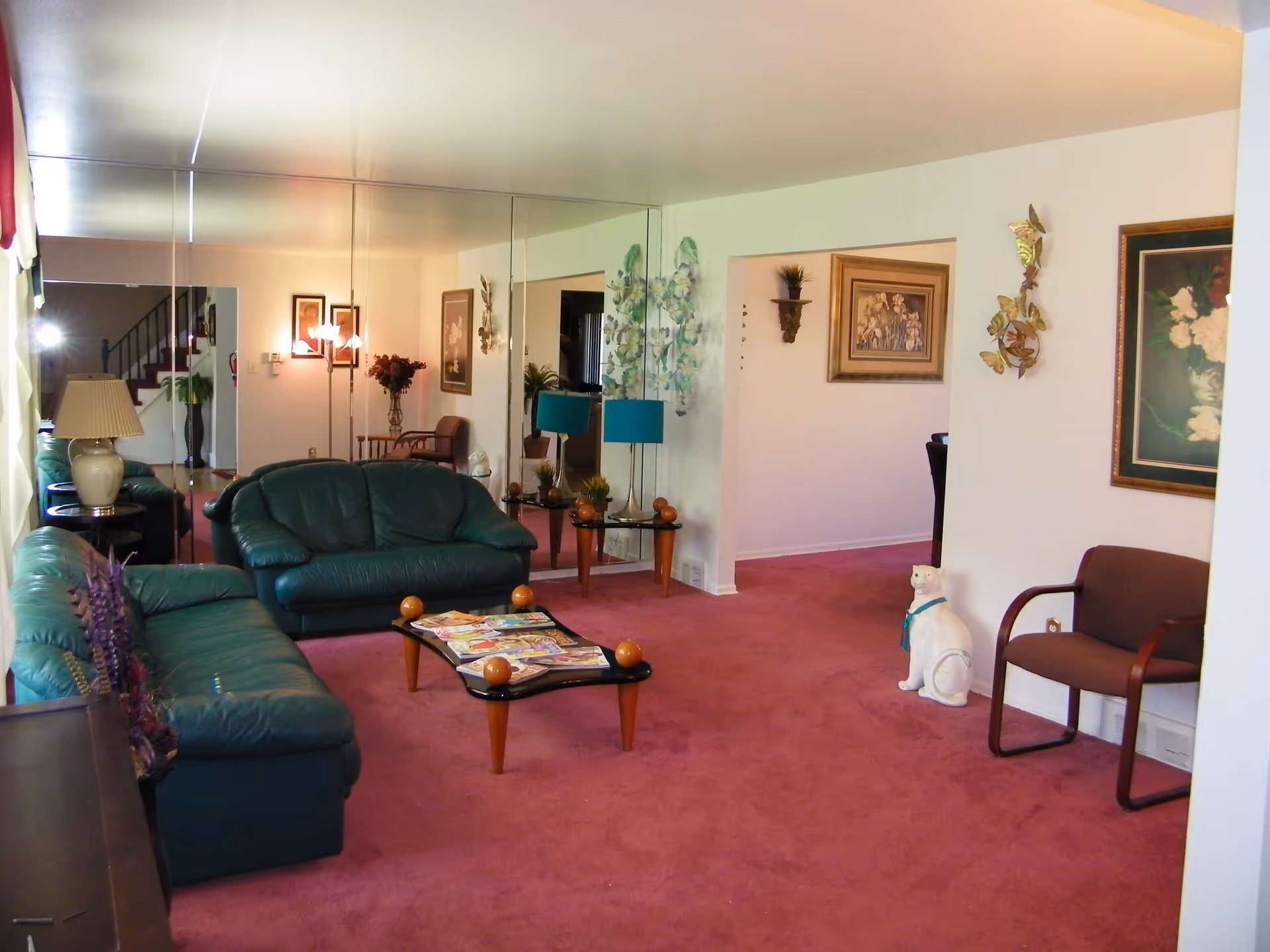 A living room with green leather sofas, a glass coffee table with magazines, a lamp, and a large wall mirror reflecting the room. The floor is covered with a pink carpet, and there are framed floral paintings and decorative items on the walls. A white ceramic cat statue with a blue ribbon sits near a brown chair.