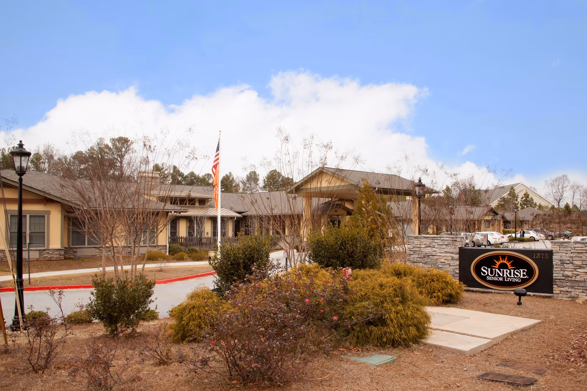 Exterior view of Sunrise Senior Living facility with a stone sign displaying the facility name, landscaped bushes and trees, a driveway, and a building under a partly cloudy sky.