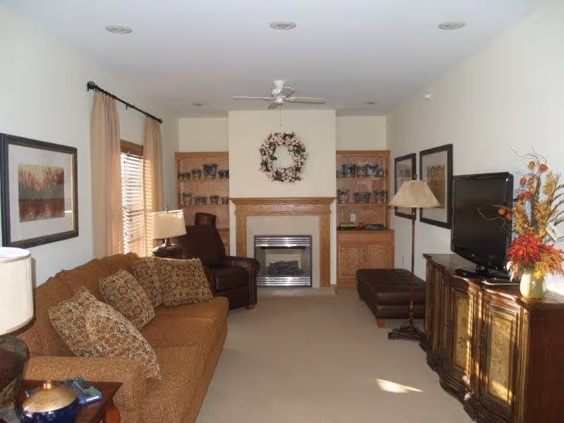 A cozy living room with a brown sofa adorned with patterned cushions on the left, a dark brown leather armchair near a window with beige curtains, a fireplace with a floral wreath above it in the center, built-in wooden shelves on either side of the fireplace, a flat-screen TV on a wooden cabinet to the right, and a floor lamp beside the TV. The walls are light-colored and the room is carpeted.