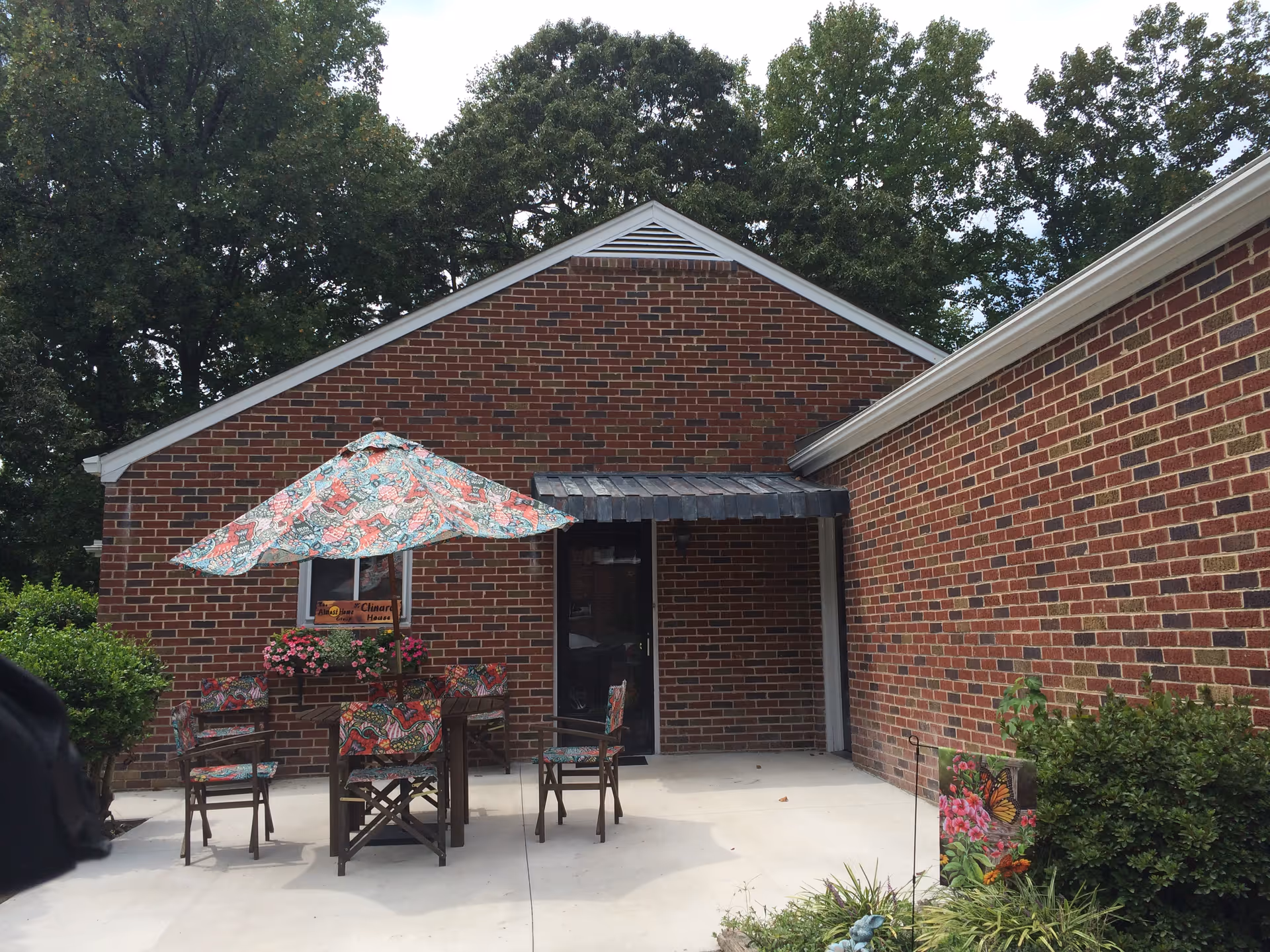 Outdoor patio area with a brick building in the background. There is a table with a colorful patterned umbrella and matching chairs. Some potted flowers are placed on the table and nearby. Green bushes and trees surround the patio.