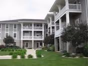 Exterior view of a multi-story senior living facility with balconies, stone and siding facade, landscaped lawn, and small trees.