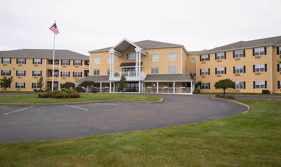 Front view of a three-story yellow senior living building with a covered entrance, circular driveway, American flag, and landscaped lawn.