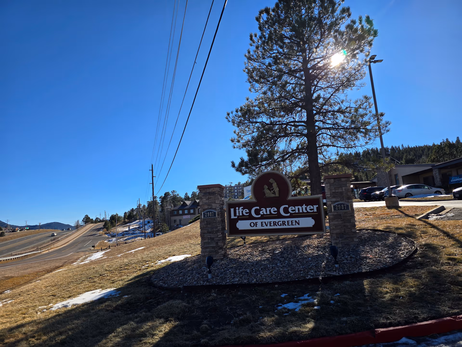 Outdoor view of the entrance sign for Life Care Center of Evergreen, situated on a grassy area with some patches of snow. The sign is mounted between two stone pillars with the address number 2987 displayed on each pillar. A tall tree and a streetlight are behind the sign, with a clear blue sky and a road running alongside the property.