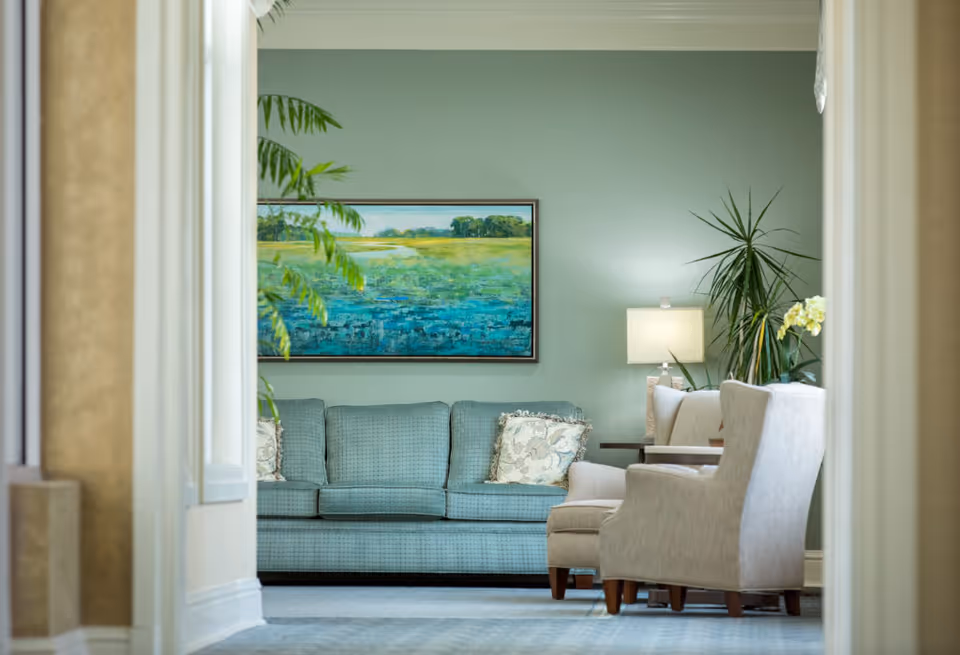View through a hallway into a living room area with a blue-green sofa adorned with patterned cushions, two beige armchairs, a side table with a lamp, and green plants. A large landscape painting with blue and green tones hangs on the wall behind the sofa.