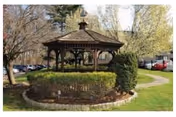 Wooden gazebo in a landscaped courtyard with shrubs, pathways, trees, and parked cars in the background.