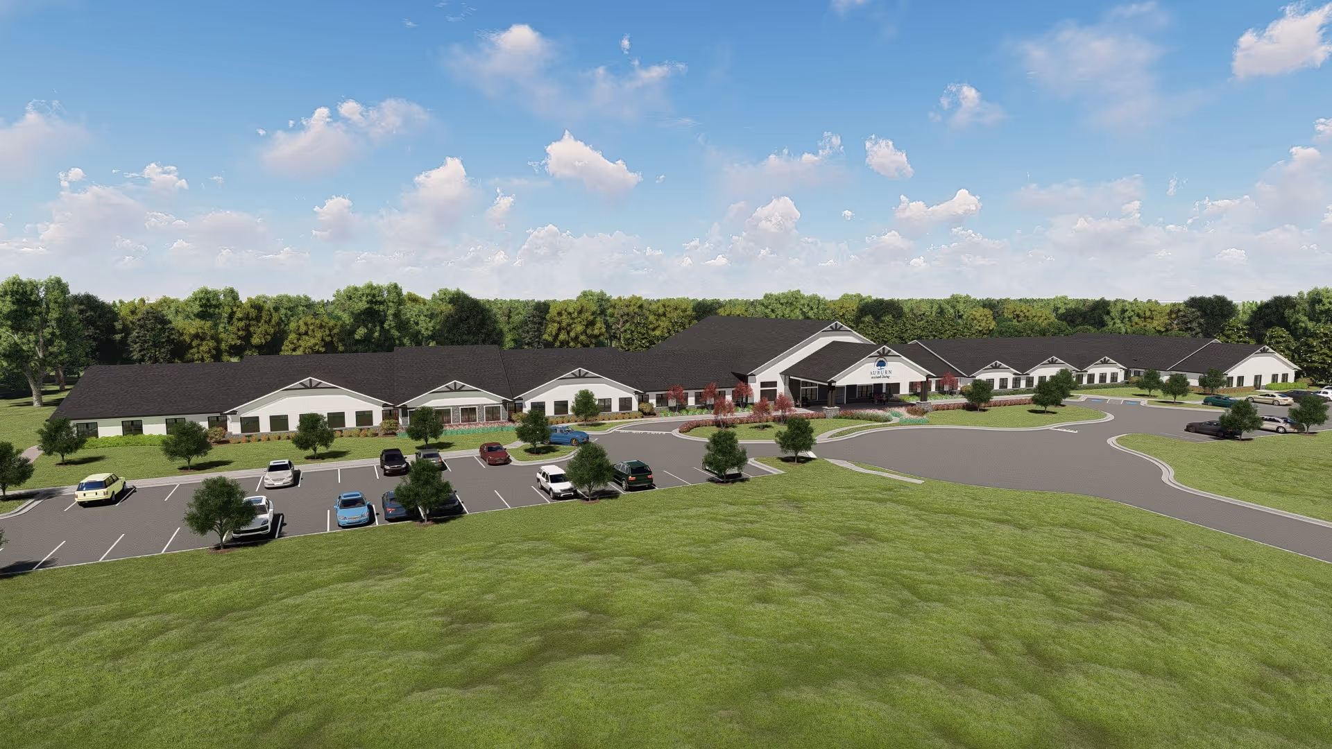 Exterior view of Auburn Assisted Living facility showing a large single-story building with a dark roof, surrounded by green lawns and trees under a blue sky with scattered clouds. There is a parking lot with several cars in front of the building.