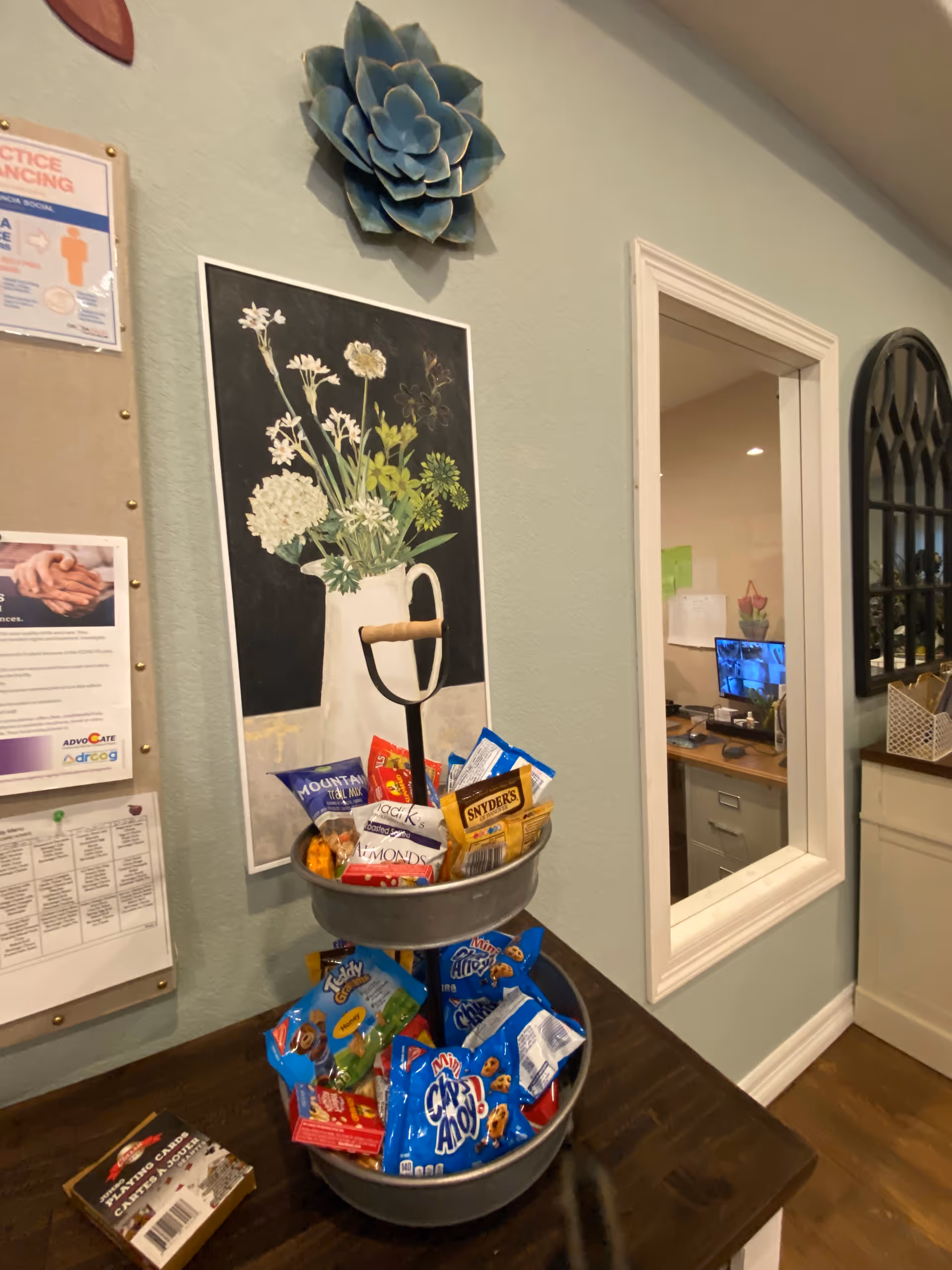 Interior reception area with a two-tiered snack display on a table beneath a floral painting and bulletin board, next to a window into an office.