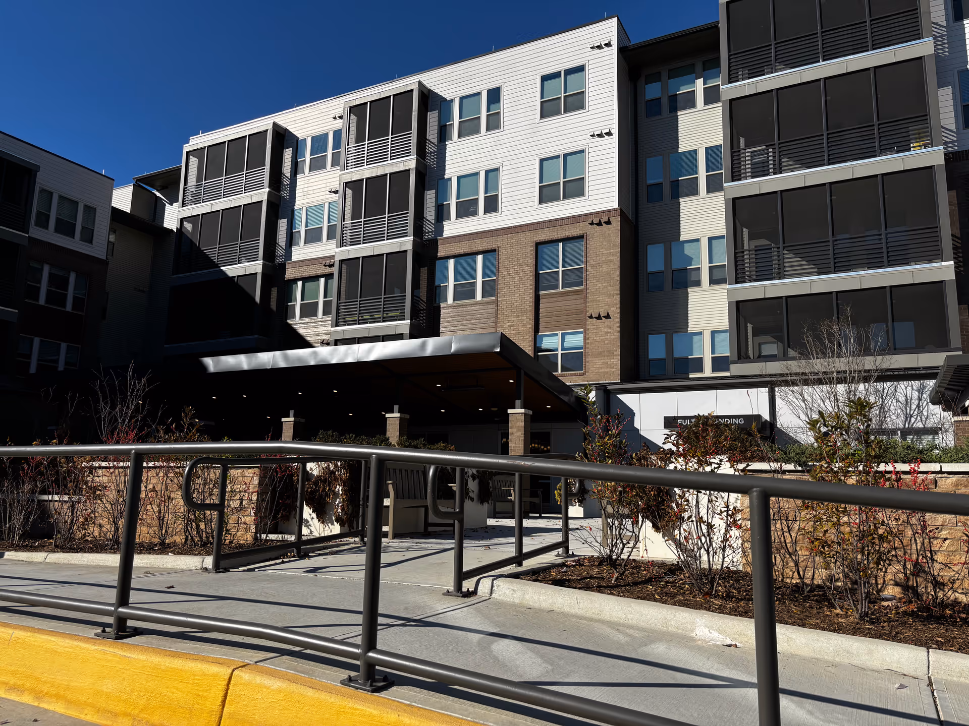 Front exterior of a modern multi-story senior living building with a covered entrance, metal railings, and landscaped planters under a clear blue sky.