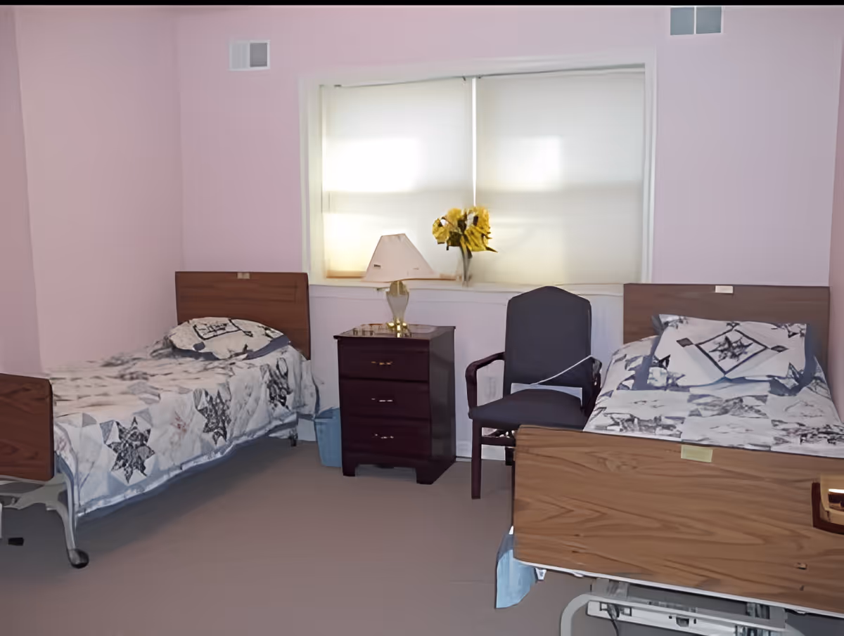 A bedroom with two hospital-style beds, each with a patterned quilt and pillow. Between the beds is a dark wooden nightstand with three drawers, a lamp, and a vase with yellow flowers. A window with closed blinds is behind the nightstand, and a dark upholstered chair is placed next to the nightstand.