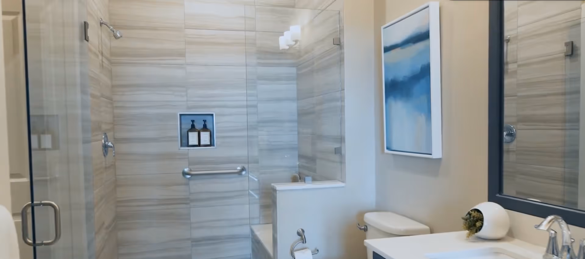 Modern bathroom with a glass-enclosed shower featuring beige and gray striped tiles, a built-in niche with two bottles, a metal grab bar, a white toilet, a white countertop with a faucet, a small plant in a white pot, a large mirror, and a blue and white abstract painting on the wall.