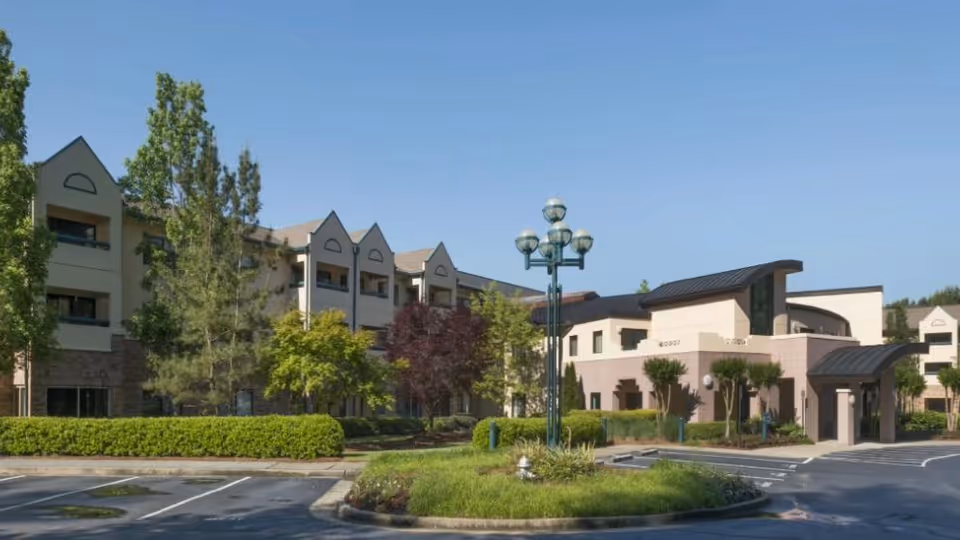 Exterior view of Delmar Gardens of Gwinnett Senior Living facility showing a multi-story building with a mix of beige and stone facade, surrounded by trees and landscaped greenery under a clear blue sky. There is a parking area and a roundabout with a decorative street lamp in the foreground.