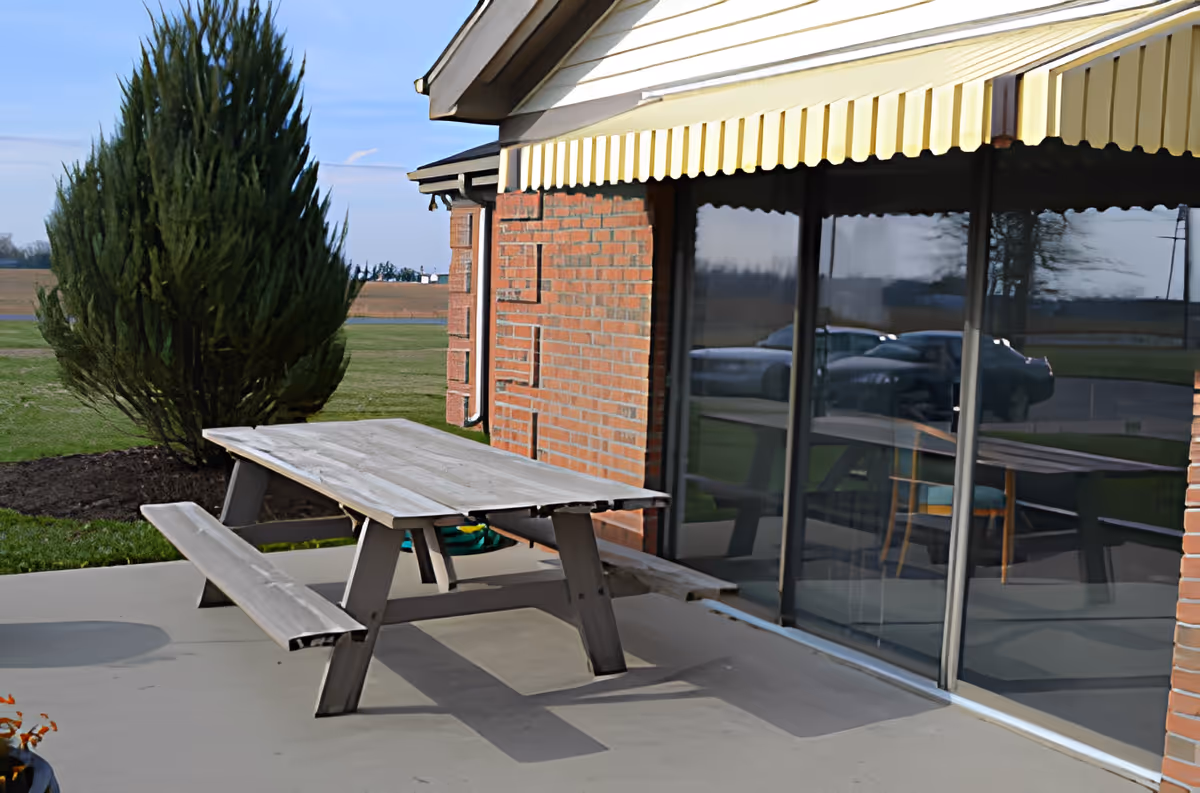 Wooden picnic table on a concrete patio beside a brick building with a yellow awning and sliding glass doors reflecting cars and the surrounding landscape.
