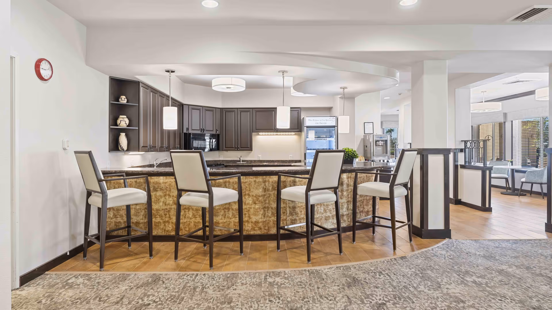 Interior view of a modern kitchen area in a senior living facility featuring a curved island with four high chairs, dark cabinetry, pendant lighting, and a refrigerator. The space is well-lit with natural light coming from windows in the adjacent dining area.