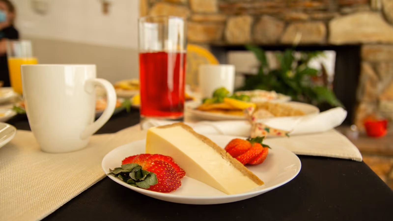 A close-up view of a dessert plate with a slice of cheesecake and fresh strawberries on a table set for a meal. In the background, there is a white coffee mug, a glass of red beverage, and other plates of food. The setting appears to be a cozy dining area with a stone fireplace and some greenery.