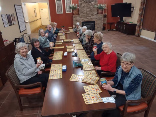 A group of elderly people sitting around a long table in a common room, holding up cans and smiling. The table is set with bingo cards and markers. The room has a stone fireplace, a large TV, and warm-colored walls.
