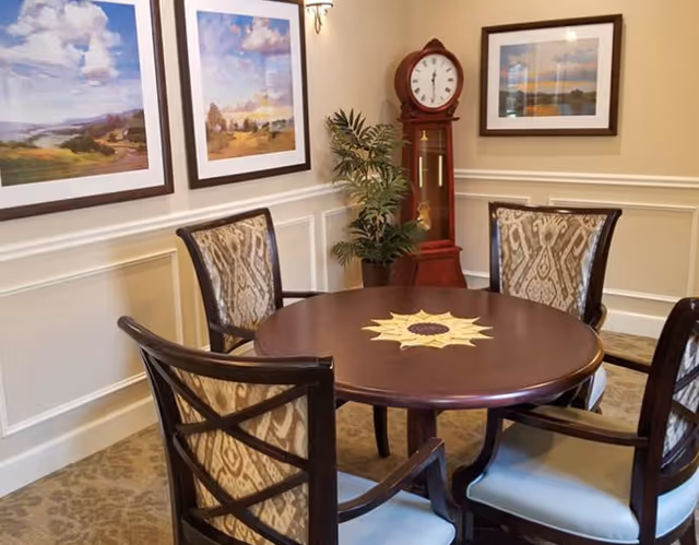 A cozy interior room with a round wooden table surrounded by four upholstered chairs. The table has a decorative sun-shaped centerpiece. On the wall behind the table, there are two framed landscape paintings and a framed photograph. A tall wooden grandfather clock and a potted plant are positioned in the corner of the room. The walls have white wainscoting and beige paint above.