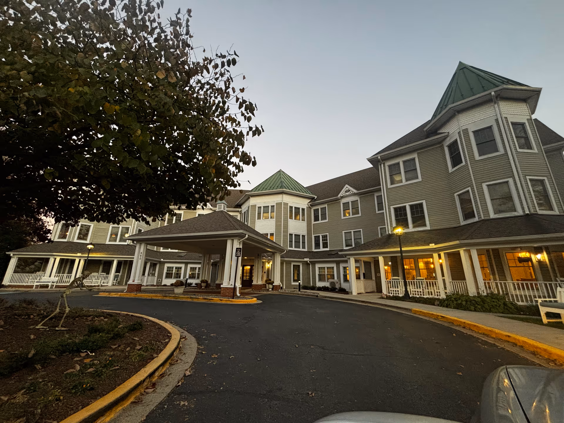 Exterior view of a multi-story senior living facility building with green roofs and multiple windows, a covered entrance driveway, and a tree on the left side during early evening.
