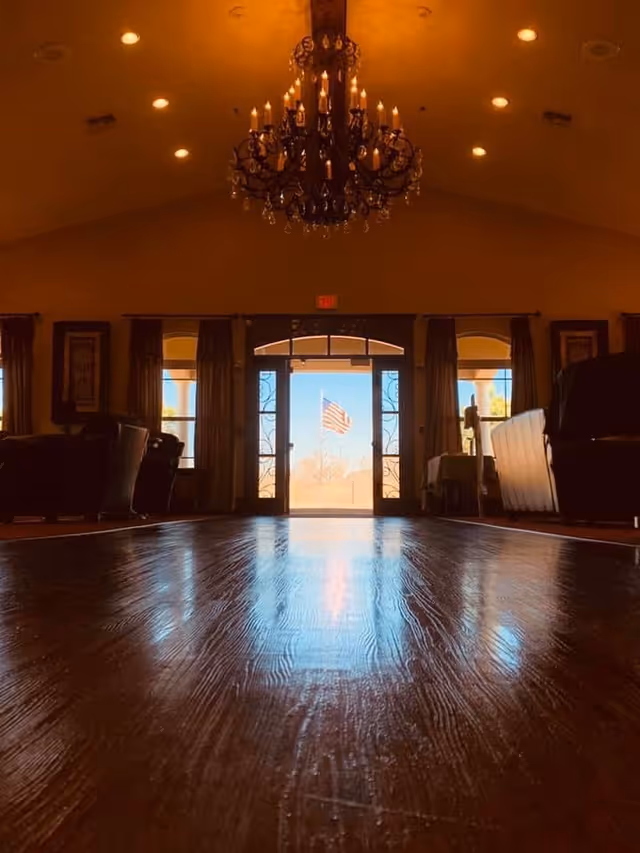 Interior view of a spacious room with polished wooden floors, a large chandelier hanging from the ceiling, and an open doorway leading outside where an American flag is visible. The room has windows with curtains and furniture along the sides.