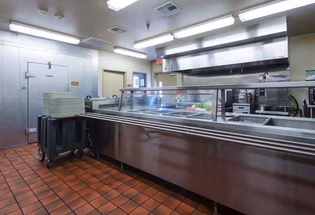 A clean commercial kitchen area with stainless steel counters and a glass sneeze guard. There are trays stacked on a cart to the left, a large walk-in refrigerator or freezer door, and various kitchen appliances and equipment in the background. The floor is covered with reddish-brown tiles and the ceiling has fluorescent lighting.