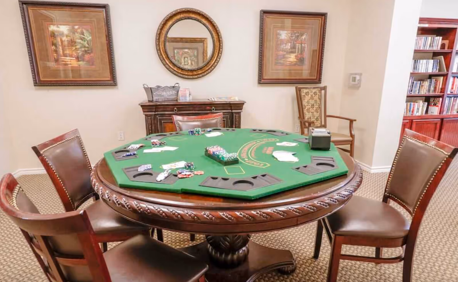 Round poker table with green felt and chips surrounded by wooden chairs in a furnished common room with artwork, a mirror, and a bookshelf.