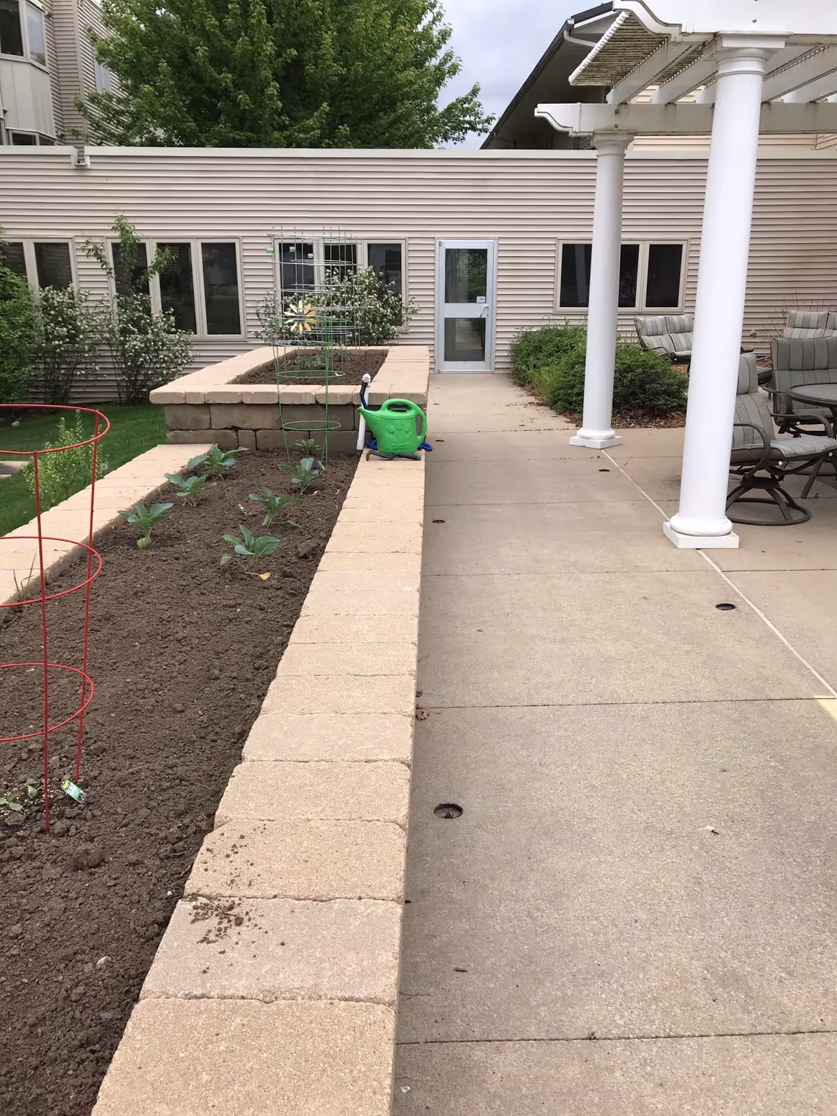 Outdoor patio area with a raised garden bed containing small plants and a green watering can. There is a concrete walkway next to the garden bed leading to a door on a beige building. On the right side, there is a covered seating area with cushioned chairs and a glass-top table. Trees and shrubs are visible in the background.