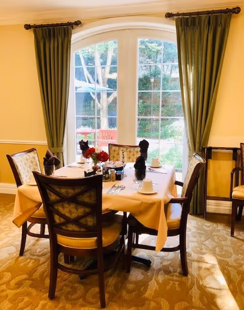 A dining table set for four with a beige tablecloth, white cups, silverware, and black napkins folded upright. The table is in front of a large window with green curtains, showing an outdoor garden area with trees and a red bench.