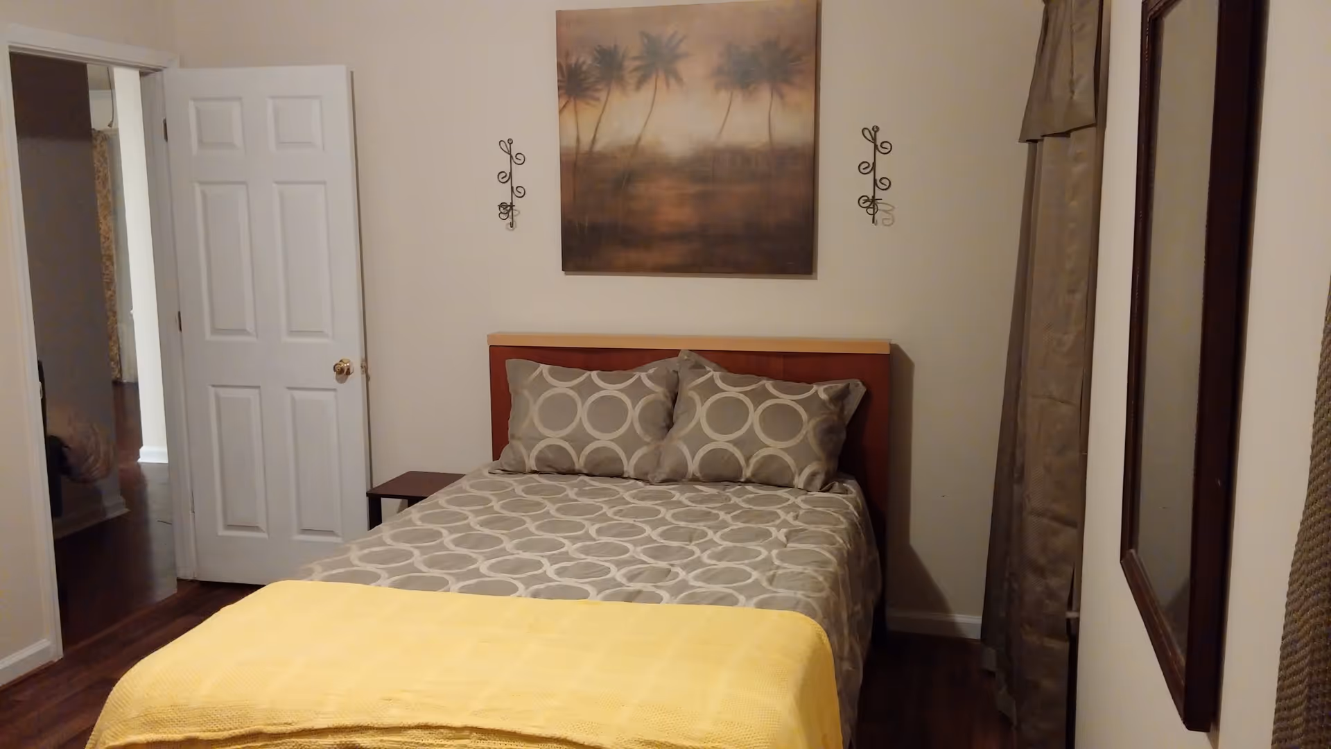 A bedroom with a bed covered in a gray and white circular patterned bedspread and two matching pillows. A yellow blanket is folded at the foot of the bed. Above the bed hangs a painting of palm trees. There are two decorative wall sconces on either side of the painting. To the left, a white door is open, showing a glimpse of another room. On the right wall, there is a window with beige curtains and a framed mirror.