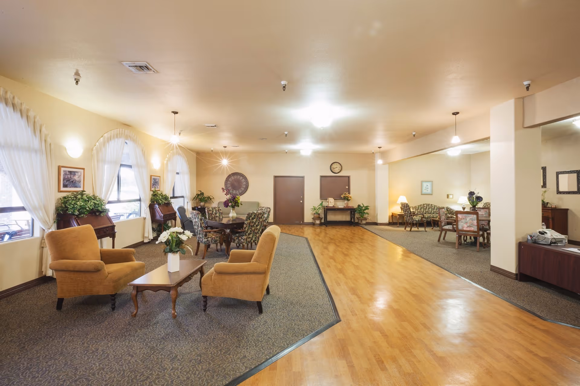 Spacious senior living common room with multiple seating areas, tables, and plants.