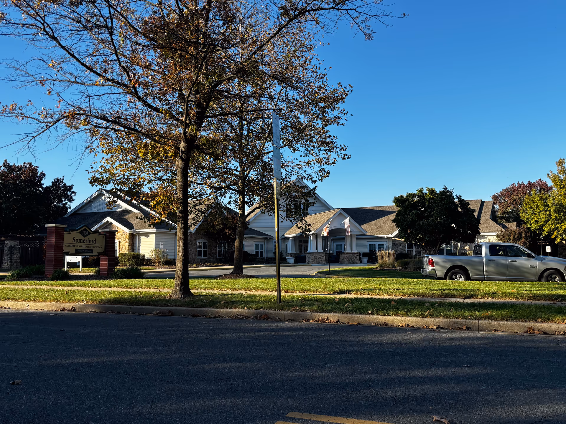 Exterior view of Somerford House Frederick, a single-story building with a pitched roof, surrounded by trees with autumn foliage. A sign in front reads 'Somerford Alzheimer's & Assisted Living Residence'. A silver pickup truck is parked on the right side of the building under a clear blue sky.