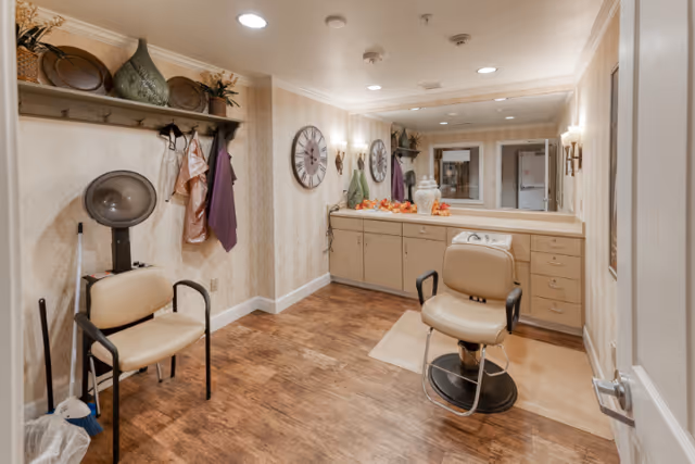 Interior of a hair salon area in a senior living facility with a salon chair, a hair dryer chair, a large mirror above a counter with cabinets, decorative items on the counter, and wall-mounted coat hooks with hanging capes and aprons.