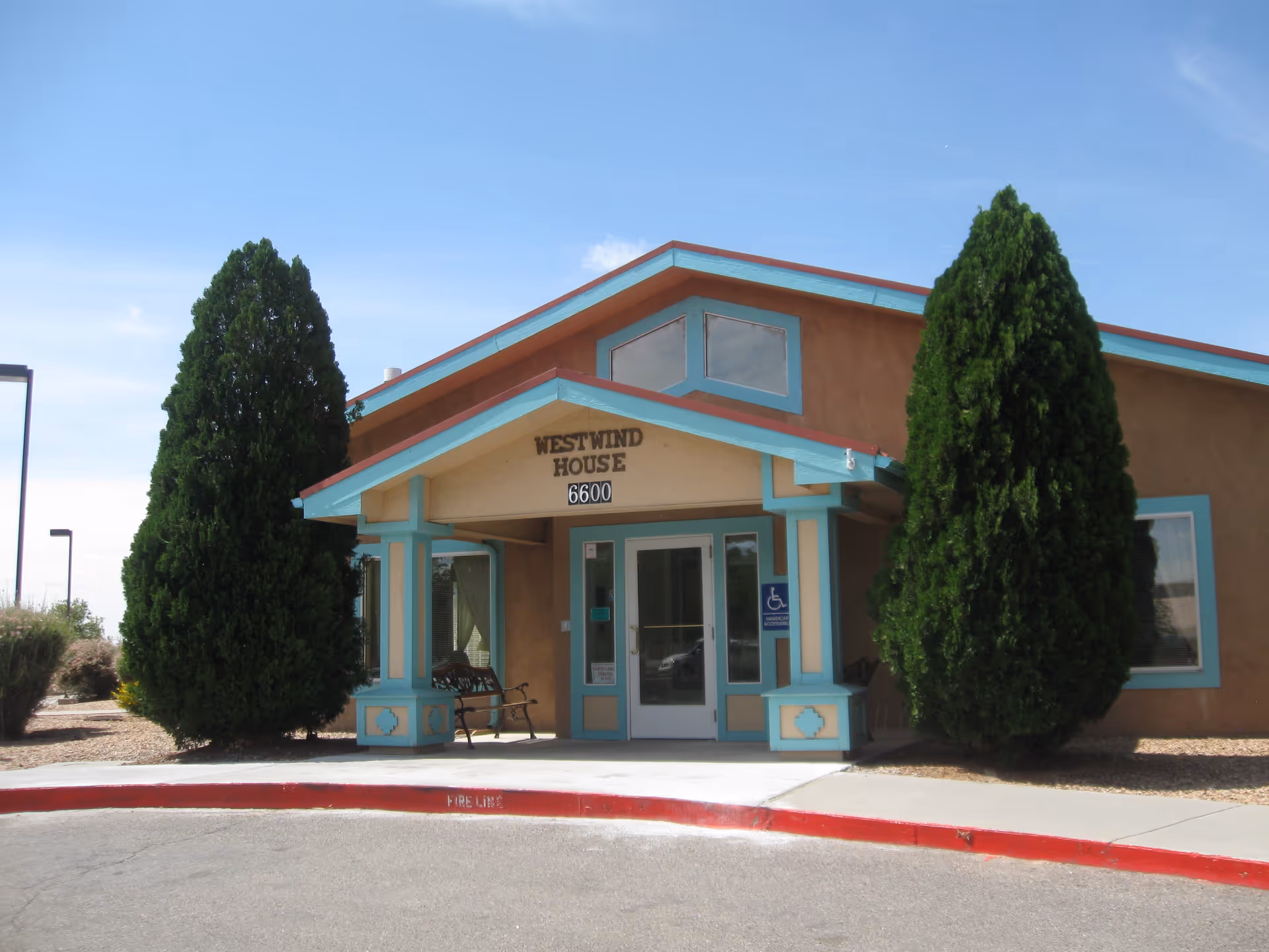 Front exterior view of Westwind House Assisted Living building with a covered entrance, two tall evergreen trees on either side, and a bench near the door. The building has a beige and turquoise color scheme and the address number 6600 is displayed above the entrance.