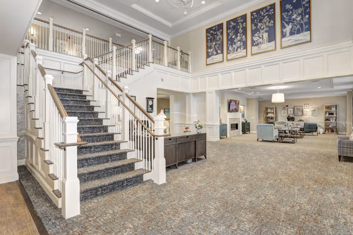 Spacious and well-lit interior of a senior living facility featuring a wide staircase with carpeted steps and white railings. The area includes a seating space with armchairs, a fireplace, bookshelves, and framed artwork on the walls. The decor is elegant with neutral tones and floral paintings above the staircase.