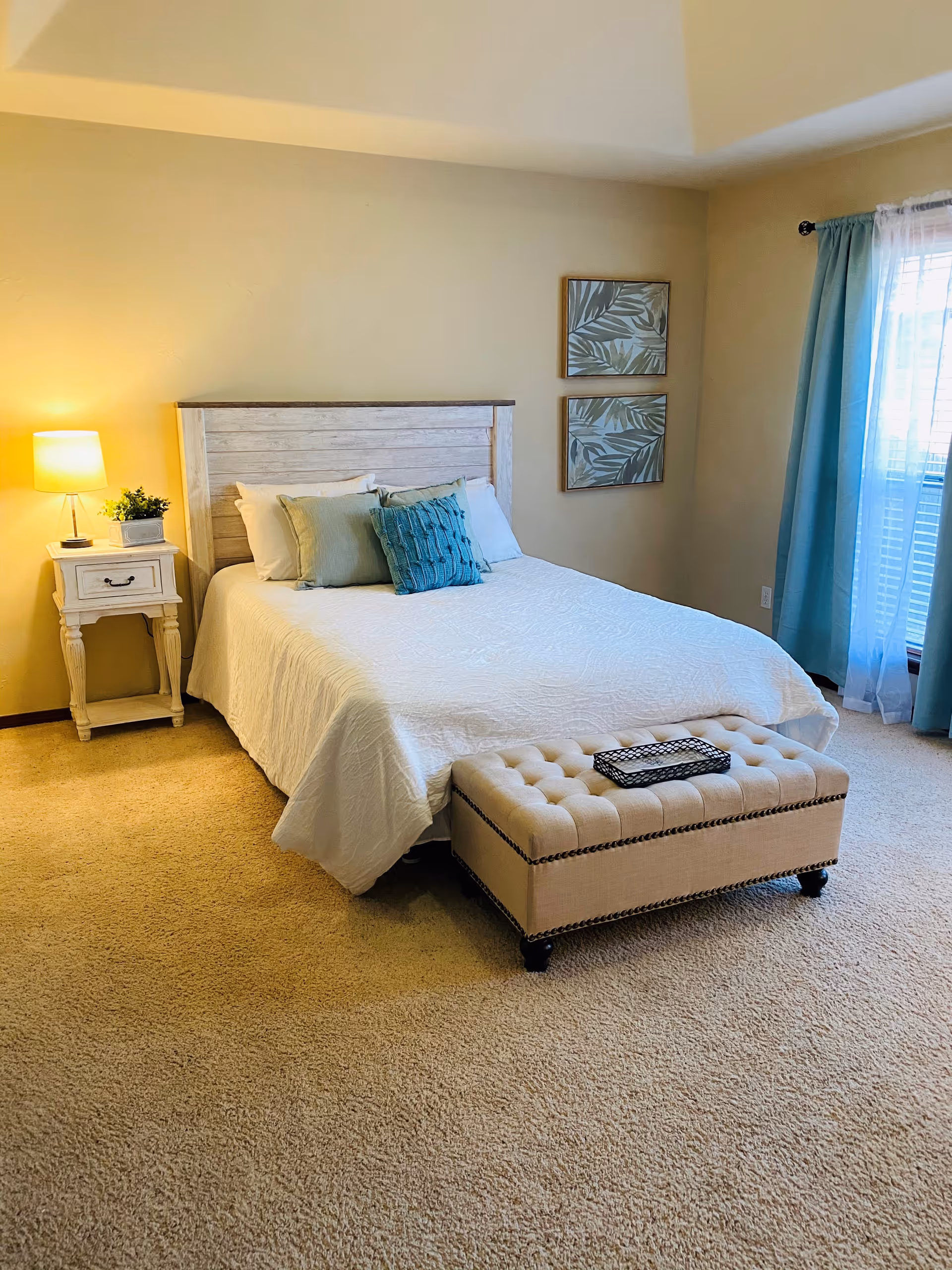 Sunlit, neutrally decorated bedroom featuring a bed with white linens and teal accent pillows, a tufted bench at the foot, a nightstand with a lamp, and a window with teal curtains.