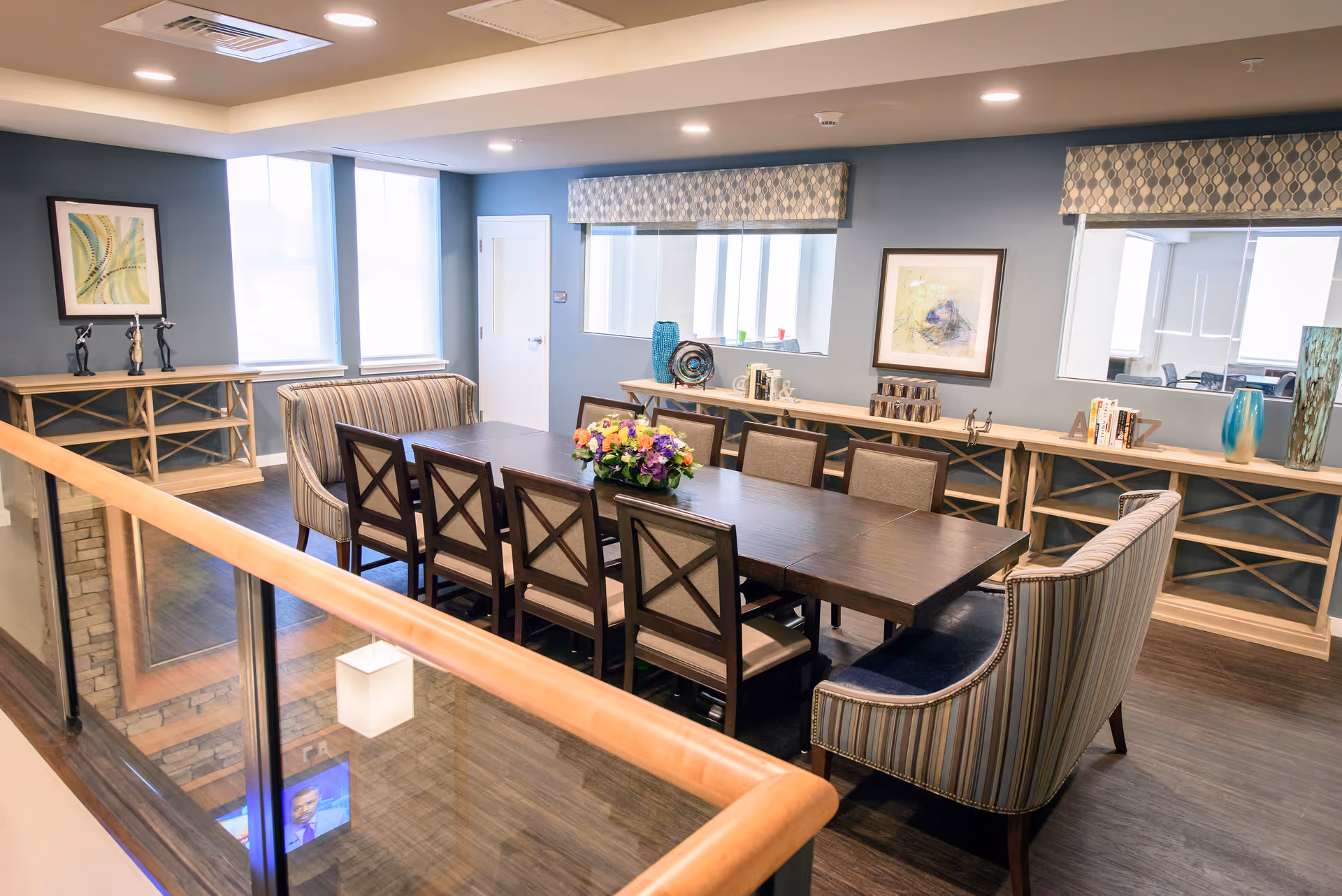 Communal dining room with a long dark wooden table surrounded by chairs and upholstered end seats, decorative shelving and artwork.