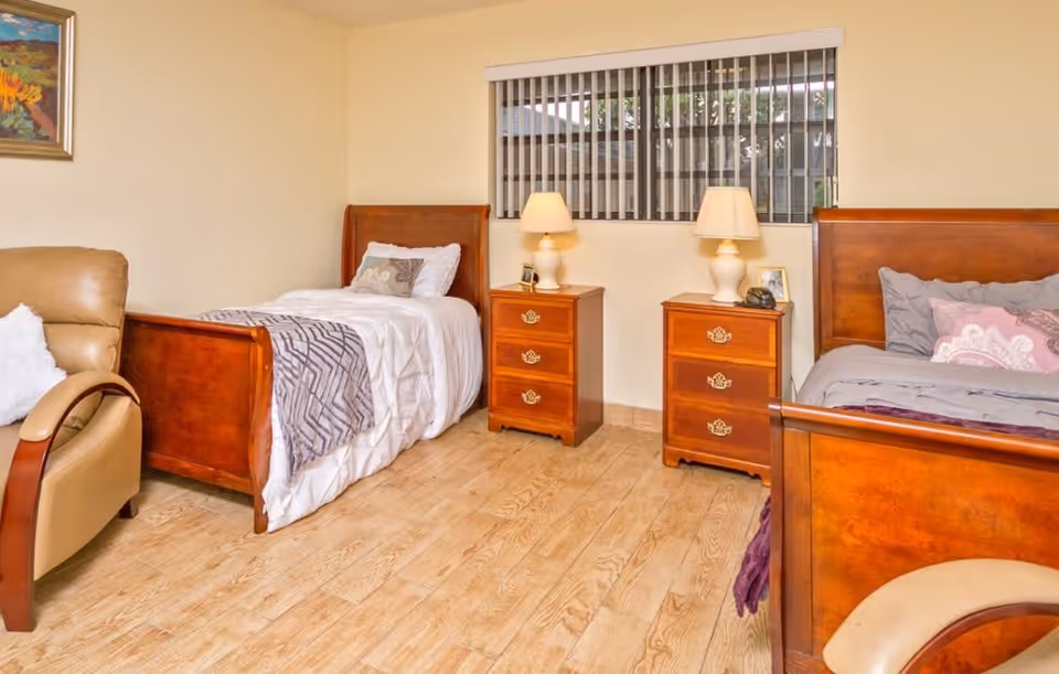 A bedroom with two wooden twin beds, each with bedding and pillows. Between the beds are two wooden nightstands, each with a lamp and small decorative items. There is a window with vertical blinds above the nightstands. A beige armchair is partially visible on the left side of the image. The floor is light wood, and the walls are painted a light cream color.
