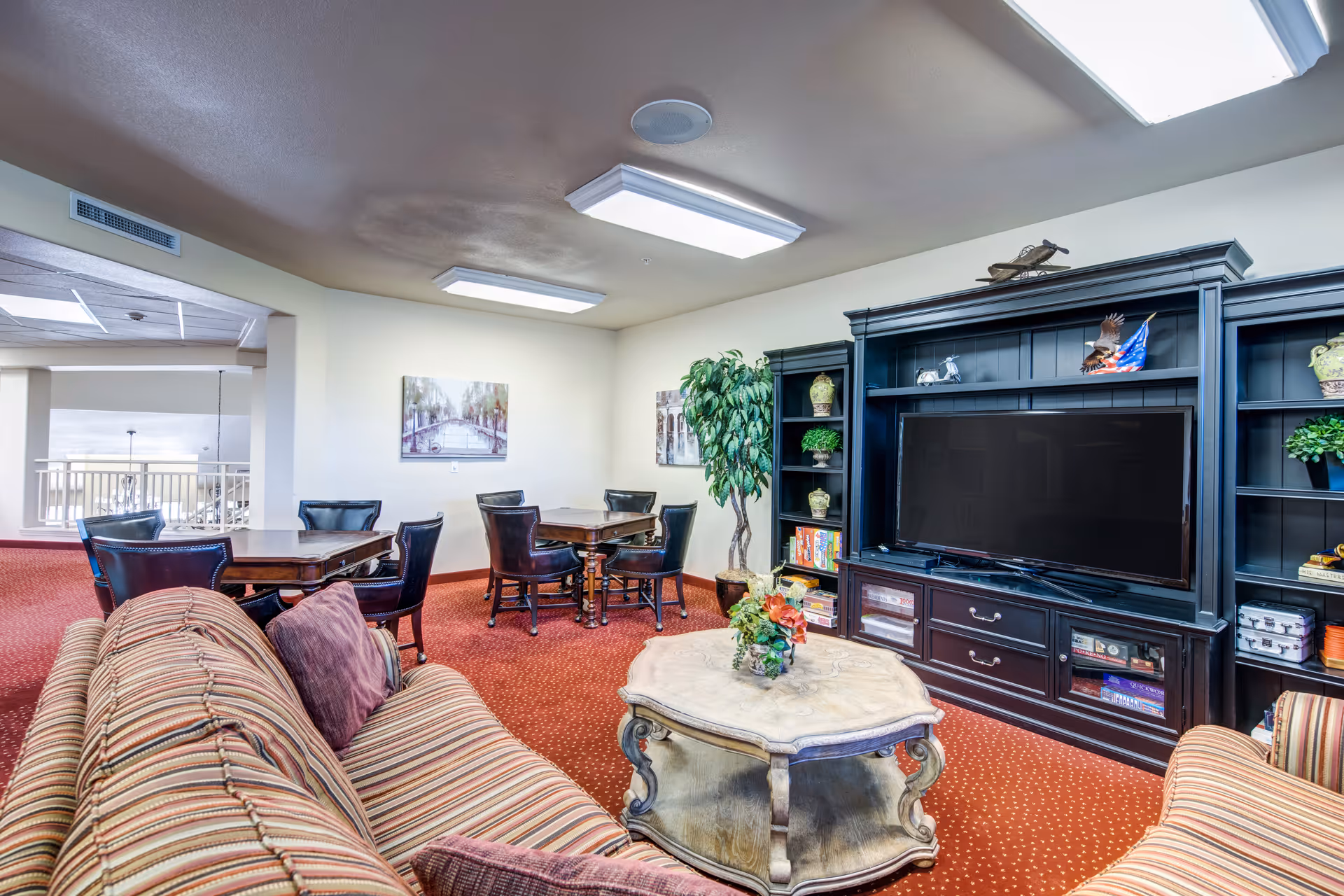 A cozy living room area with a striped sofa and a matching armchair facing a large black entertainment center with a flat-screen TV. The entertainment center has shelves with decorative items and books. In front of the sofa is a wooden coffee table with a flower arrangement. In the background, there are two wooden tables each surrounded by four chairs, and two framed pictures on the wall. The room has red carpet flooring and bright ceiling lights.