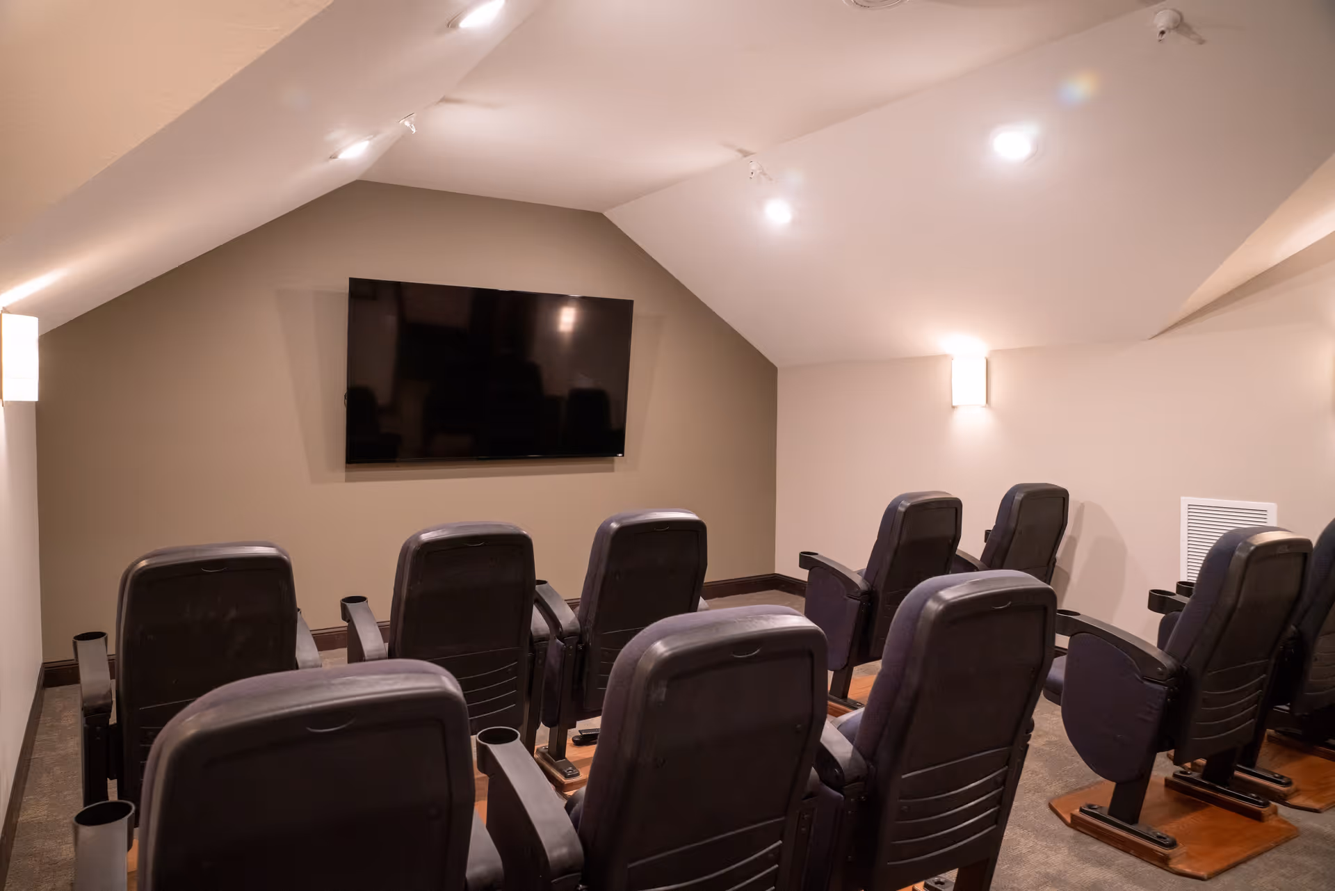 Small theater room with two rows of black cushioned chairs facing a large flat screen TV mounted on a beige wall. The room has slanted ceilings with recessed lighting and wall sconces providing soft illumination.