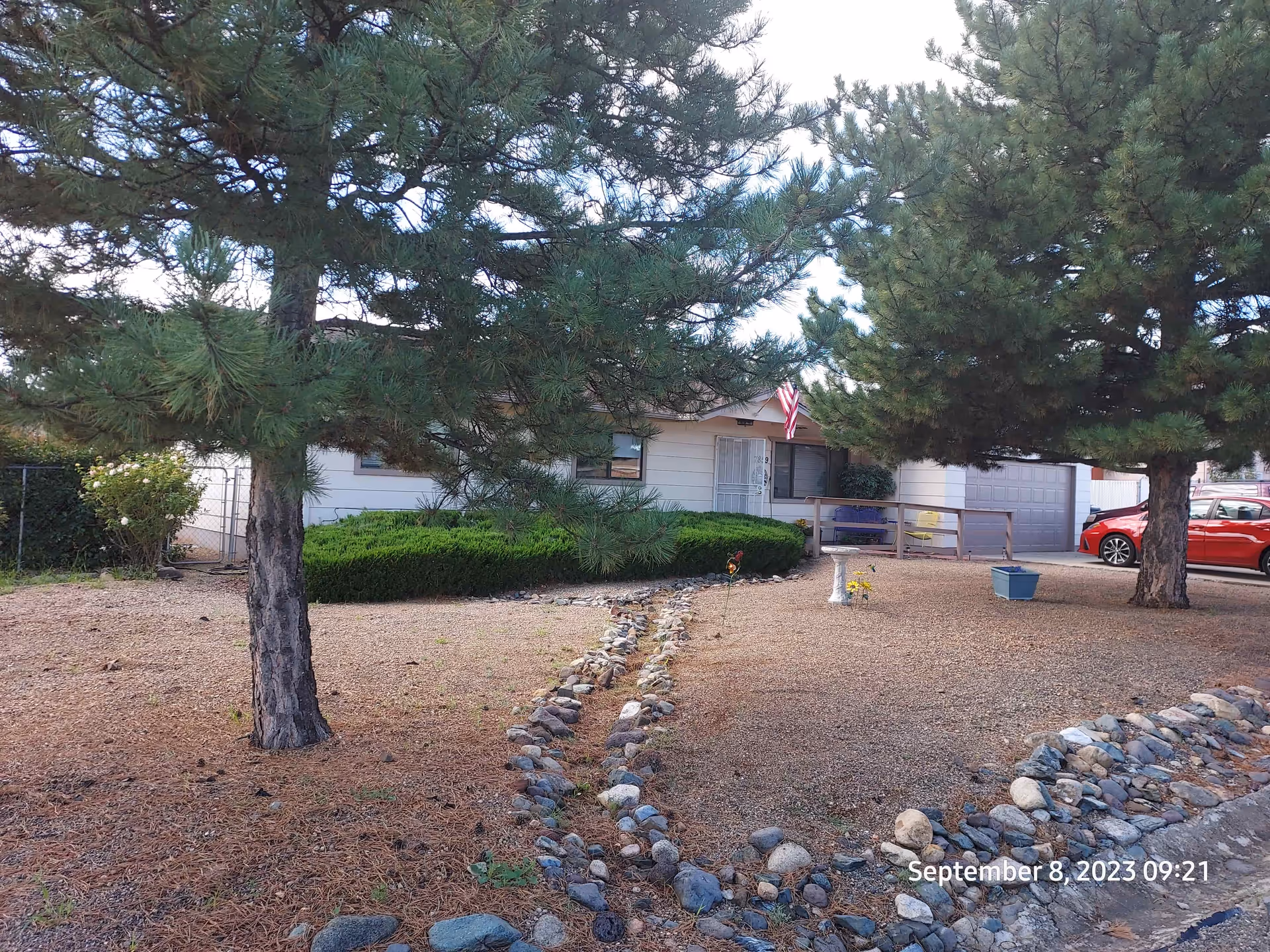 Front yard of a single-story house with two large pine trees, a small garden with bushes, a stone-lined dry creek bed, and an American flag near the entrance. A red car is parked near the garage on the right side. The ground is covered with dry pine needles and gravel.